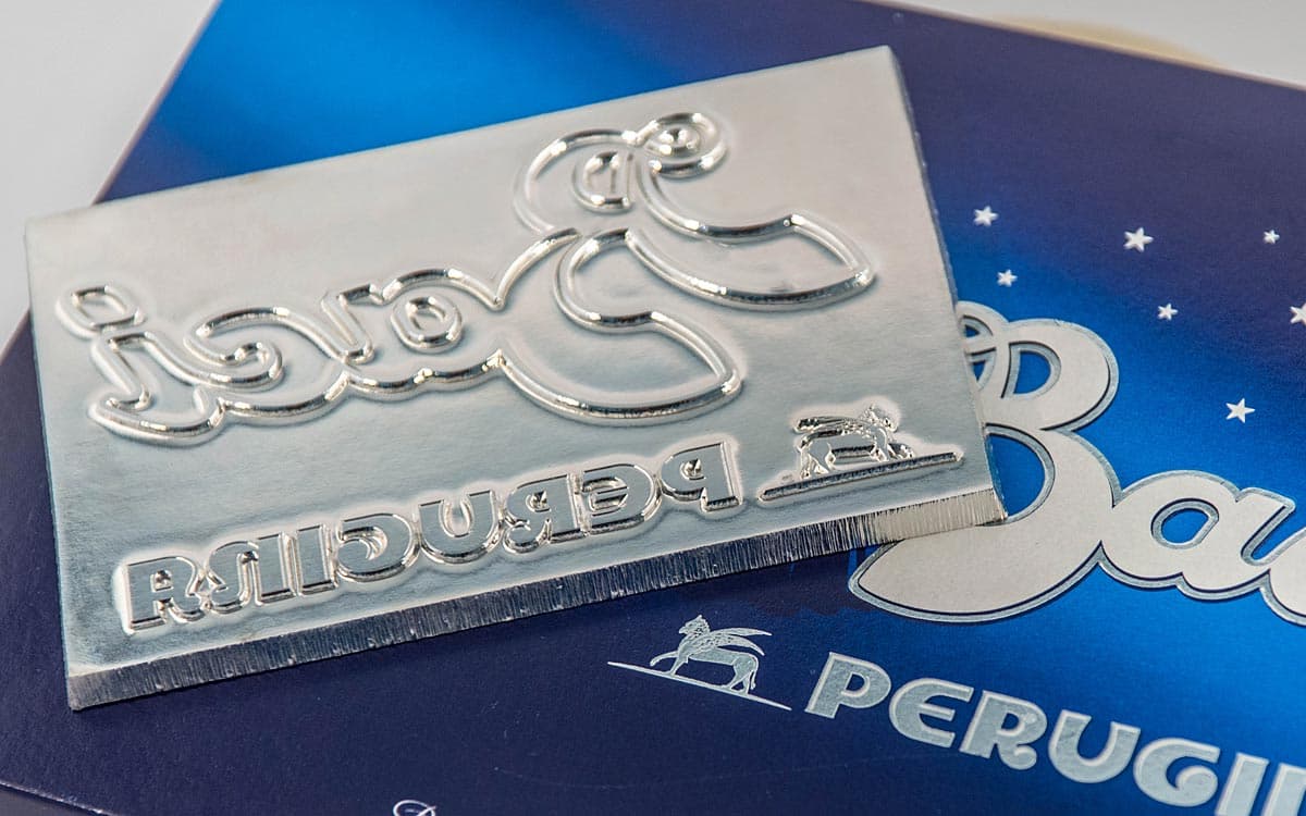 Matrice metallica con logo Baci Perugina specchiato appoggiata su una scatola di cioccolatini blu.